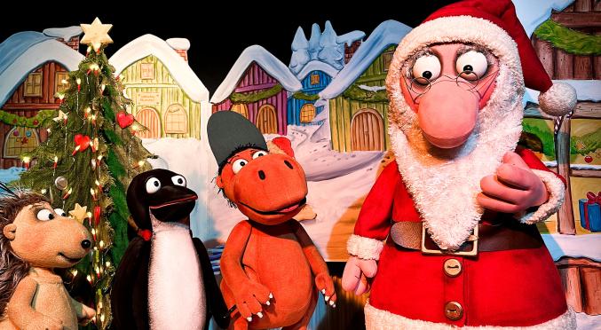 Weihnachtsszene: Santa Claus mit Renten, Pinguin, Igel und Nilpferd in Kostümen vor beleuchtetem Weihnachtsbaum und bunten Häusern.
