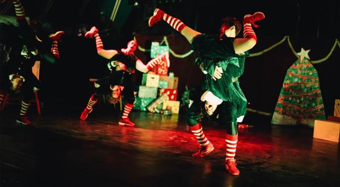 Tänzer in grünen und schwarzen Kostümen mit rot-weißen Strümpfen und Schuhen, dynamische Posen auf dunkler Bühne mit Weihnachtsbaum.