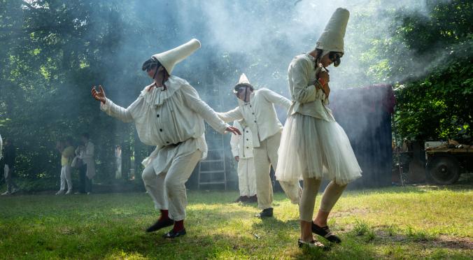 Vier Figuren in weißen Pulcinella-Kostümen mit großen Hüten und Masken, drei tanzen auf Waldwiese im Sonnenlicht mit Nebel, eine im Hintergrund.