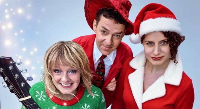 Drei Musiker in Weihnachtskleidung: blonde Frau mit Schneeflocken-Pullover und Gitarre, Mann in rotem Anzug mit Federhut, Frau im Santa-Kleid mit Keyboard, hellblauer Hintergrund.