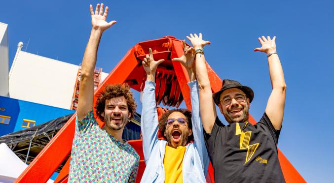 Drei Personen mit erhobenen Armen vor großer roter Metallstruktur: gemustertes Hemd, gelbe Jacke mit Sonnenbrille, schwarzes T-Shirt mit Blitz, blauer Himmel.