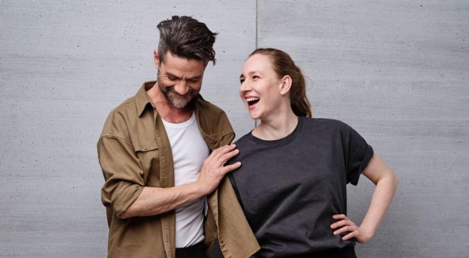 Ein Mann und eine Frau stehen vor einer grauen Wand. Beide lachen, der Mann trägt ein khakifarbenes Hemd, die Frau ein graues T-Shirt.