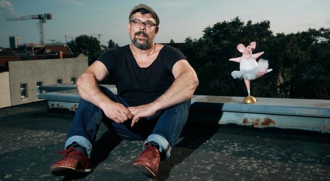 Ein Mann sitzt auf einem Dach und trägt ein schwarzes T-Shirt, Jeans und rote Schuhe. Neben ihm steht eine kleine, rosa Ballerina-Figur.