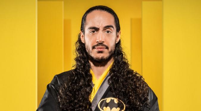Ein Mann mit langen lockigen Haaren trägt ein schwarzes Oberteil mit Batman-Logo und steht vor einem gelben Hintergrund.