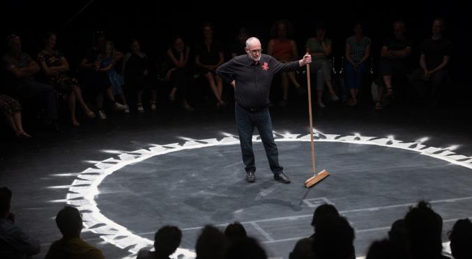  Ein älterer Mann steht in einem dunklen Theaterraum im Zentrum eines Kreises aus weißem Pulver und hält einen Besen, während das Publikum im Hintergrund sitzt.