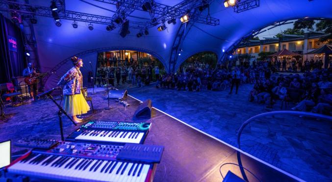 Eine Sängerin mit buntem Outfit steht barfuß auf einer Open-Air-Bühne unter einem Zelt, umgeben von Musikern. Ein großes Publikum sitzt im Halbrund, während blaue Lichter die Szene erhellen.