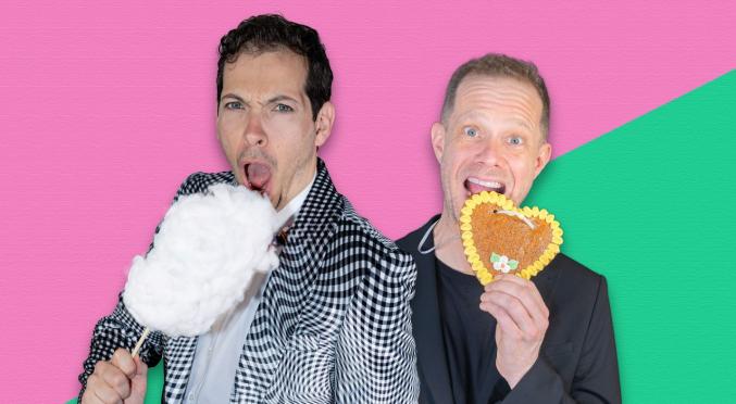 Zwei Männer vor einem farbigen Hintergrund, einer mit Zuckerwatte, der andere mit einem Lebkuchenherz, beide lächeln in die Kamera.