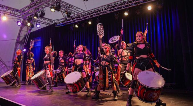 Das Foto zeigt eine Gruppe von Musikern auf einer Bühne. Die Musiker tragen markante, auffällige Kostüme in Schwarz und Rot. Einige von ihnen spielen große Trommeln, die sie vor ihrem Körper tragen, während andere verschiedene Perkussionsinstrumente halten. Sie alle wirken energetisch und engagiert, und einige heben ihre Instrumente oder Trommelstöcke in die Luft. Die Beleuchtung der Bühne ist dramatisch, mit bunten Lichtern, die die Szenerie hervorheben. 