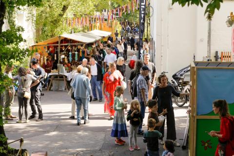  Auf dem Bild ist eine lebhafte Szene bei einem Straßenfest oder Markt zu sehen. Viele Menschen, darunter Erwachsene und Kinder, bewegen sich durch eine belebte Gasse. Im Hintergrund sind Marktstände mit bunten Stoffen und Dekorationen. Über der Szene hängen bunte Girlanden oder Fähnchen.