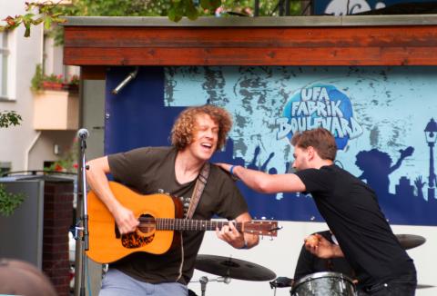  Auf dem Bild ist eine musikalische Live-Performance zu sehen. Im Vordergrund spielt ein Mann mit lockigen Haaren leidenschaftlich eine Akustikgitarre. Neben ihm ist ein weiterer Mann, der sich offensichtlich in einer Interaktion mit dem Gitarristen befindet