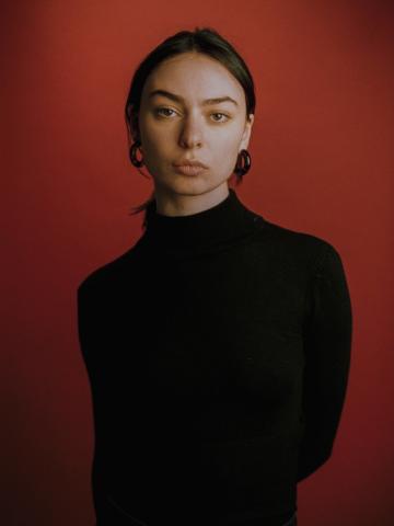 Ein Porträt der Künstlerin Salome Topuria vor einem dunkelroten Hintergrund. Sie blickt direkt in die Kamera, ihr Ausdruck wirkt selbstbewusst und künstlerisch. Der warme, rote Hintergrund unterstreicht ihre Präsenz und verleiht dem Bild eine intensive, ausdrucksstarke Atmosphäre.