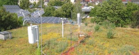 Auf dem begrünten Flachdach der ufaFabrik sind diverse meteorologische Messinstrumente installiert. Die weißen Gehäuse der Geräte heben sich deutlich vom satten Grün der Vegetation ab. Der Blick auf die Stadt im Hintergrund unterstreicht die städtische Nutzung des nachhaltig gestalteten Gebäudes.