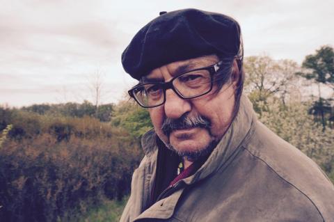 Mann mit Brille, Hut und Bart blickt ernst, trägt Mantel und steht vor einer herbstlichen Landschaft.