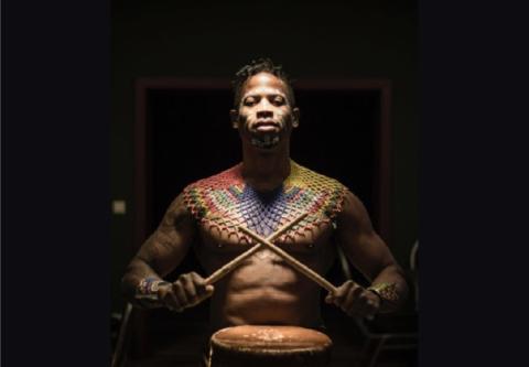Das Bild zeigt einen Musiker, der an einem Drum sitzt. Er trägt eine farbenfrohe, kunstvolle Halskette und hat einen konzentrierten Ausdruck im Gesicht. Der Hintergrund ist dunkel, was die Details des Musikers und seiner Kleidung hervorhebt. Die Atmosphäre ist kraftvoll und ausdrucksstark.