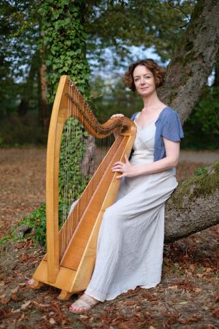 Eine Frau mit lockigem Haar sitzt an einem Baumstamm und spielt eine hölzerne Harfe. Sie trägt ein langes, helles Kleid mit einer blauen Jacke. Der Hintergrund ist grün und herbstlich.