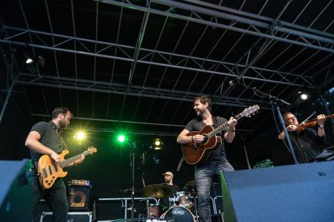 ine Band spielt auf einer Open-Air-Bühne mit schwarzem Dach. Ein Gitarrist lächelt, während er spielt, daneben ein Bassist, ein Schlagzeuger und ein Geiger. Bunte Scheinwerfer leuchten.