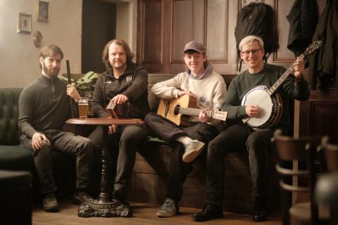 Das Bild zeigt vier Männer, die in einem gemütlichen Raum mit Holzmöbeln sitzen. Sie halten Instrumente: eine Flöte, eine Bodhrán, eine Gitarre und ein Banjo. Die Szene vermittelt eine entspannte und freundliche Atmosphäre, die typisch für ein informelles Musikkreis- oder Pub-Setting ist.