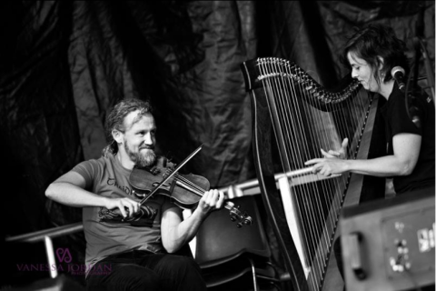 Das Bild zeigt zwei Musiker auf einer Bühne, die intensiv miteinander spielen. Einer spielt Geige, während die andere an einer Harfe sitzt. Die Aufnahme ist in Schwarz-Weiß gehalten, was die Emotionen und die Dynamik der Darbietung verstärkt. Der Hintergrund ist dunkel, wodurch die Musiker im Fokus stehen. Copy Regenerate