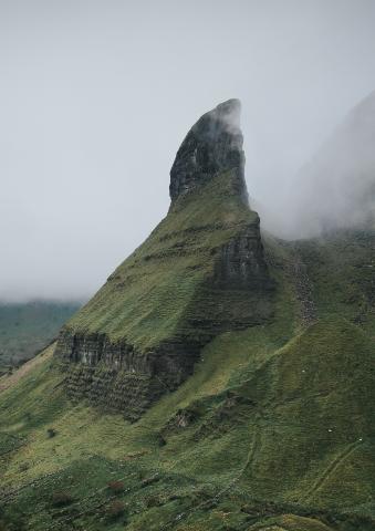 Ein markanter, grasbewachsener Berg ragt spitz in einen nebligen Himmel. Die steilen Hänge sind teils felsig und von Nebel umhüllt, was eine mystische Atmosphäre schafft.