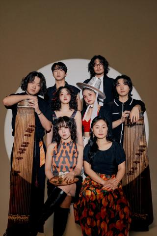 Acht junge Menschen in stilvoller Kleidung posieren vor kreisförmigem Licht. Zwei halten traditionelle koreanische Instrumente, eine trägt einen Hut im Hanbok-Stil.