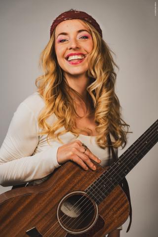 Eine junge Frau mit lockigem Blondhaar lächelt breit, trägt ein weißes Shirt und hält eine Gitarre. Sie wirkt fröhlich und leidenschaftlich beim Musizieren.