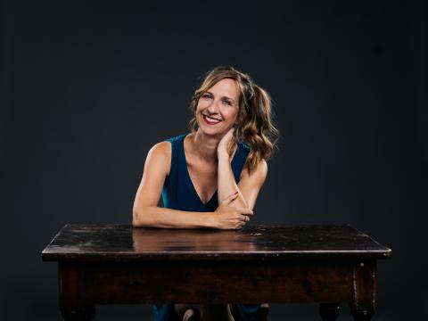 Eine Frau mit lockigem Haar sitzt freundlich an einem dunklen Tisch, lächelt, trägt ein blaues Top und wirkt entspannt vor dunklem Hintergrund.