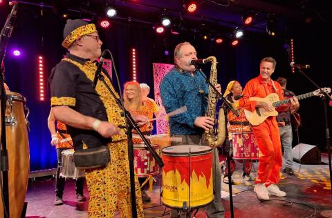 Eine bunte Band spielt auf der Bühne. Musiker tragen farbenfrohe Kleidung, spielen Saxophon, Gitarre und Trommeln. Im Hintergrund tanzen und trommeln weitere Mitglieder unter buntem Licht.