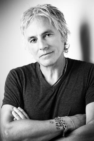 Porträt eines Mannes mit hellen, zurückgekämmten Haaren, dunklem T-Shirt und Armketten, verschränkten Armen, vor hellem Hintergrund in Schwarz-Weiß.