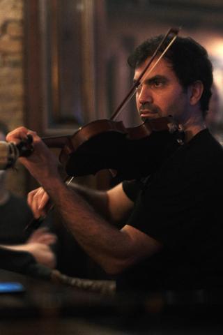 Konzentrierter Musiker mit dunklem Haar spielt Violine, hält den Bogen in der rechten Hand, trägt schwarzes Hemd, im Hintergrund unscharf ein Klavier.