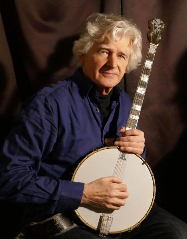 Älterer Mann mit grauen Haaren hält ein Banjo, trägt ein blaues Hemd, sitzt lächelnd vor dunklem Hintergrund, Instrument mit Resonator und Markierung ‚Fair Quality‘.