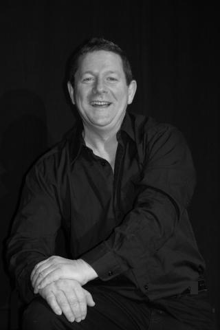 Lächelnder Mann mit kurzen Haaren, dunklem Hemd und aufgestützten Händen, sitzend vor schwarzem Hintergrund in Schwarz-Weiß-Fotografie.