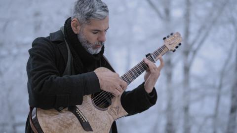 Mann mit graumeliertem Bart und schwarzem Schal spielt konzentriert eine hellbraune Akustikgitarre im Winterwald mit kahlen Bäumen.