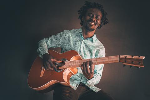 Lächelnder Mann mit Dreadlocks und gemustertem Hemd spielt eine braune Akustikgitarre, sitzt entspannt vor dunklem Hintergrund.