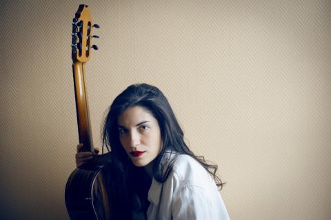 Frau mit langen dunklen Haaren und rotem Lippenstift hält eine klassische Gitarre vor hellbrauner, gemusterter Tapete, Blick leicht seitlich in die Kamera.