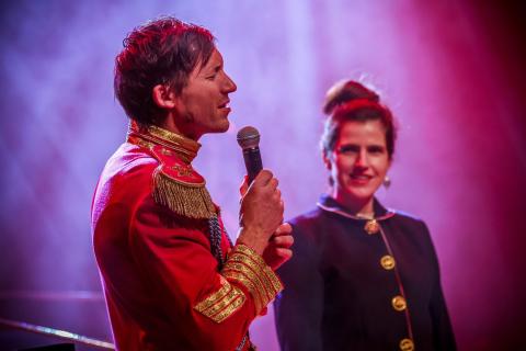 Mann in roter Uniform mit goldenen Epauletten hält ein Mikrofon, spricht lächelnd auf der Bühne; im Hintergrund eine lächelnde Frau mit hochgesteckten Haaren in dunkler Jacke.