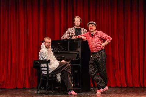 Drei Männer auf einer Bühne vor rotem Vorhang: Einer sitzt mit weißem Pelzkragen am Klavier, einer lehnt im karierten Hemd am Klavier, einer steht mit Clownnase in rotem Hemd und grauer Hose.