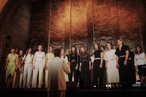Chor aus 14 Personen in hellen und dunklen Outfits steht singend auf einer Bühne mit Ziegelsteinmauer-Hintergrund, eine Person dirigiert im Vordergrund.