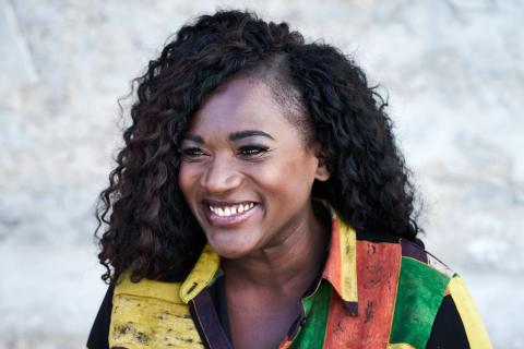 Lächelnde Person mit lockigem Haar, seitlich rasiert, trägt ein buntes Hemd in Gelb, Grün und Orange, vor hellem, unscharfem Hintergrund.