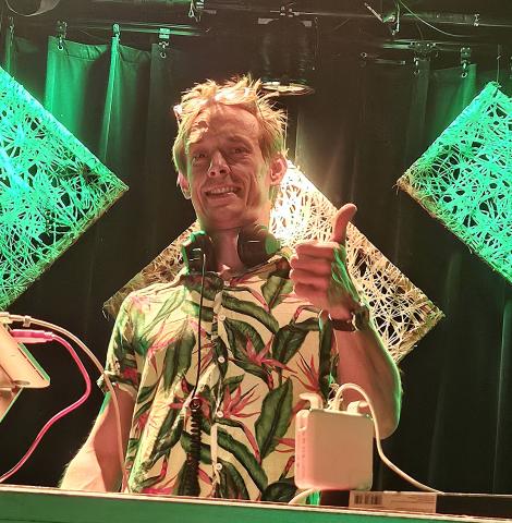 Lächelnder Mann mit blonden Haaren in blumigem Hemd steht auf der Bühne, gibt Daumen hoch, Mikrofon in der Hand, grüne LED-Dekoration im Hintergrund.