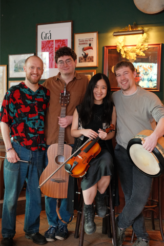 Vier lächelnde Personen in gemütlicher Kneipe: Zwei halten Gitarre und Rahmentrommel, eine hält eine Violine, im Hintergrund Bilder und Guinness-Deko, entspannte und freundliche Atmosphäre.