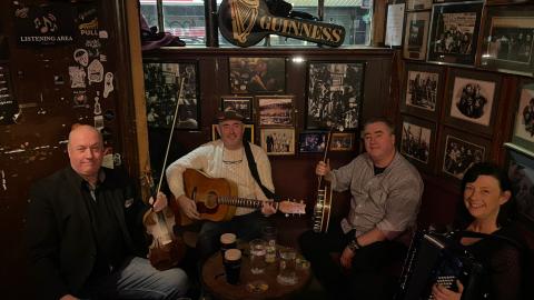 Vier lächelnde Personen in gemütlicher Kneipe: Zwei halten Musikinstrumente (Gitarre, Akkordeon), Tisch mit Gläsern, im Hintergrund Fotos, Guinness-Schilder und irische Deko.