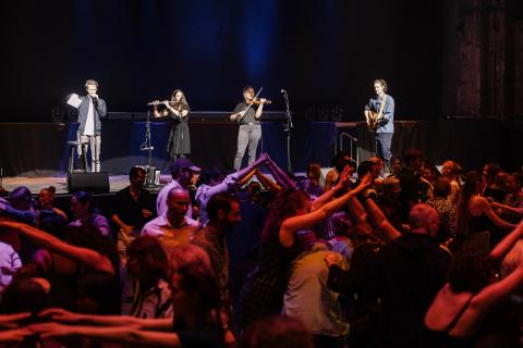 Vier Musiker:innen auf der Bühne: Links Person mit Mikrofon, Mitte zwei mit Flöte und Violine, rechts Person mit Gitarre, jubelndes Publikum mit erhobenen Armen, blaue Bühnenbeleuchtung.