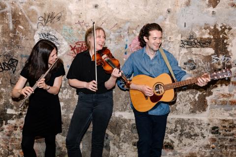 Drei lächelnde Musiker:innen vor Graffiti-Wand: Links Person mit Querflöte, Mitte Person mit Violine, rechts Person mit Gitarre, alle in lässiger Kleidung, lebendige und fröhliche Atmosphäre.