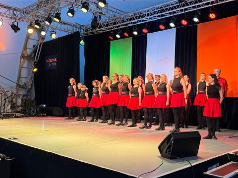 Gruppe von 14 Tänzer:innen auf der Bühne in schwarzer Oberbekleidung und roten Röcken, stehend in einer Reihe, Bühne mit Scheinwerfern und ufaFabrik-Berlin-Banner im Hintergrund.