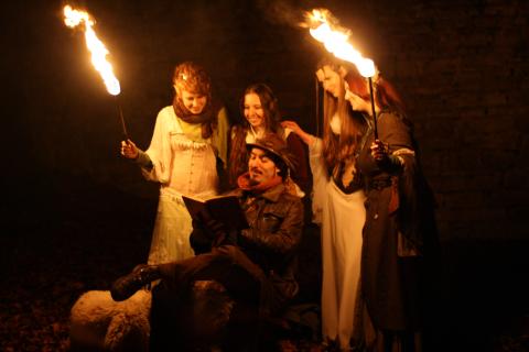 Fünf Personen in mittelalterlicher Kleidung bei Nacht: Eine liest aus einem Buch, vier halten brennende Fackeln, alle lächelnd, mystische und gemütliche Atmosphäre.