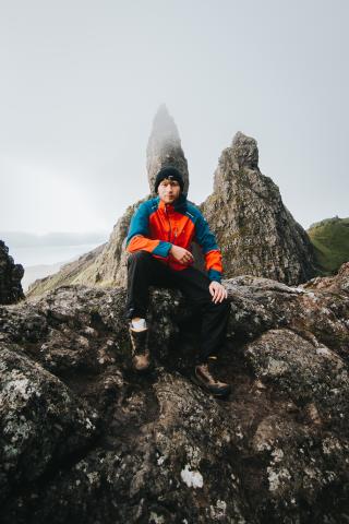 Person sitzt auf felsigem Gipfel mit spitzen Felsformationen im Hintergrund, trägt blaue-orange Wanderjacke, schwarze Hose und Mütze, bewölkter Himmel, weite Landschaft.