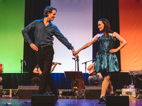 Zwei lächelnde Personen tanzen auf der Bühne: Mann in dunklem Hemd und Hose, Frau in glänzendem, türkisfarbenem Kleid, halten sich an den Händen, im Hintergrund Musiker:innen mit Instrumenten.