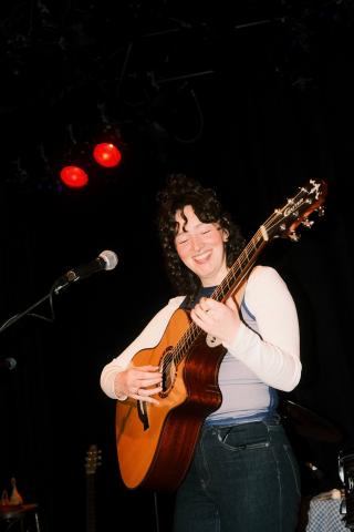 Lächelnde Person spielt Gitarre auf der Bühne, Mikrofon im Hintergrund, rote Lichter, dunkle Kleidung mit hellem Langarm-Shirt, geschlossene Augen, entspannte und fröhliche Atmosphäre.