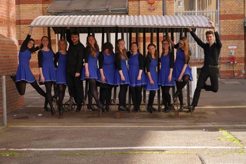 Neun lächelnde Personen posieren unter einem Metall-Vordach: Acht in blauen Kleidern mit schwarzen Strumpfhosen, einer in schwarzer Kleidung, alle halten das Dach fest, dynamische und fröhliche Gruppenstimmung.