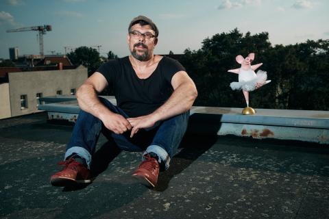 Ein Mann sitzt auf einem Dach und trägt ein schwarzes T-Shirt, Jeans und rote Schuhe. Neben ihm steht eine kleine, rosa Ballerina-Figur.